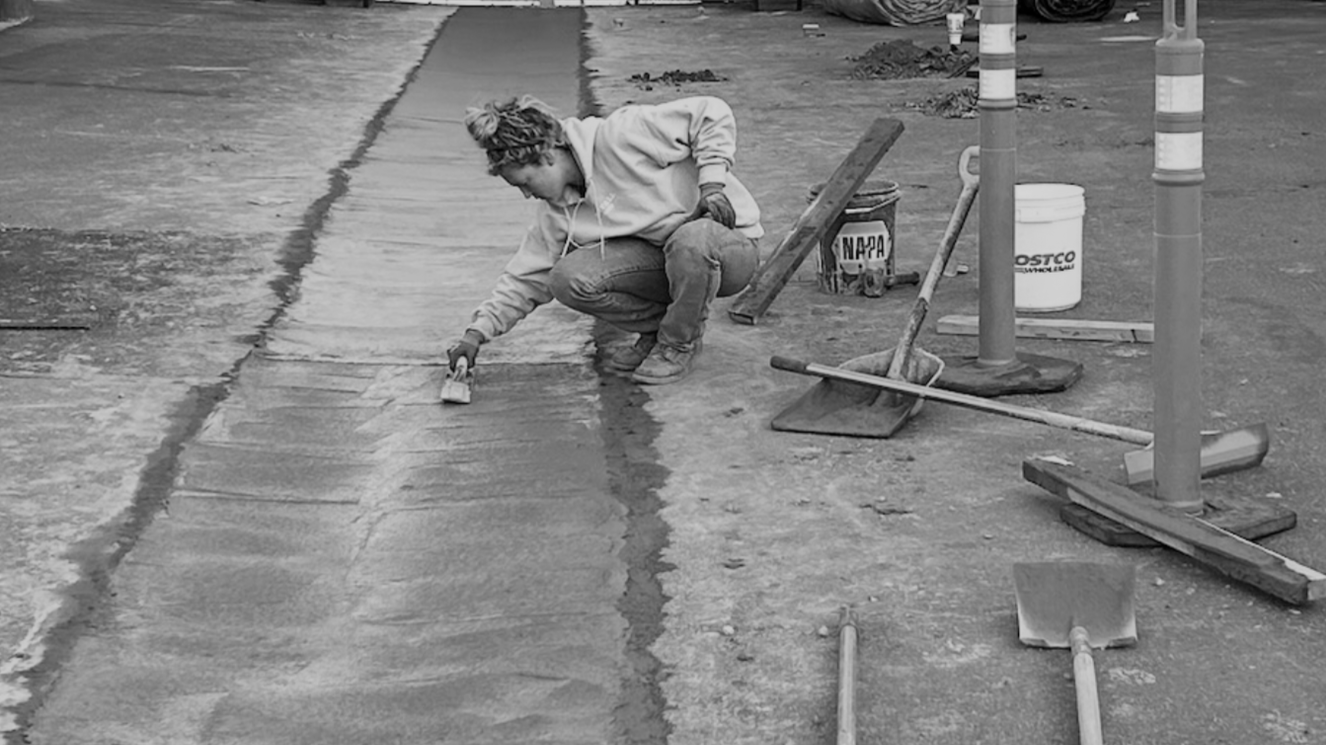 A woman working on a concrete sidewalk, using a trowel to smooth the surface, surrounded by construction tools and safety cones.