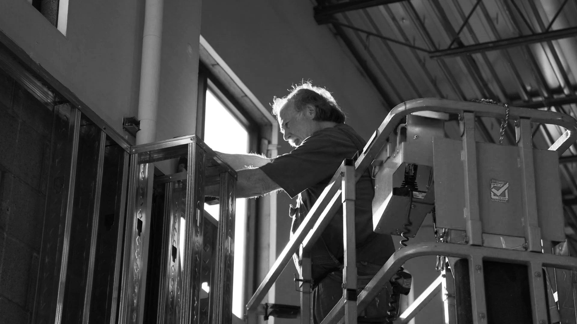 A worker on a lift installing electrical or communication wiring in a building's ceiling, with natural light coming through a window.