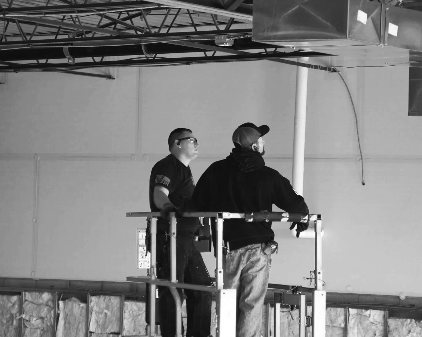 Two workers on a scissor lift working on the ceiling of a building, with visible HVAC ductwork and wiring.