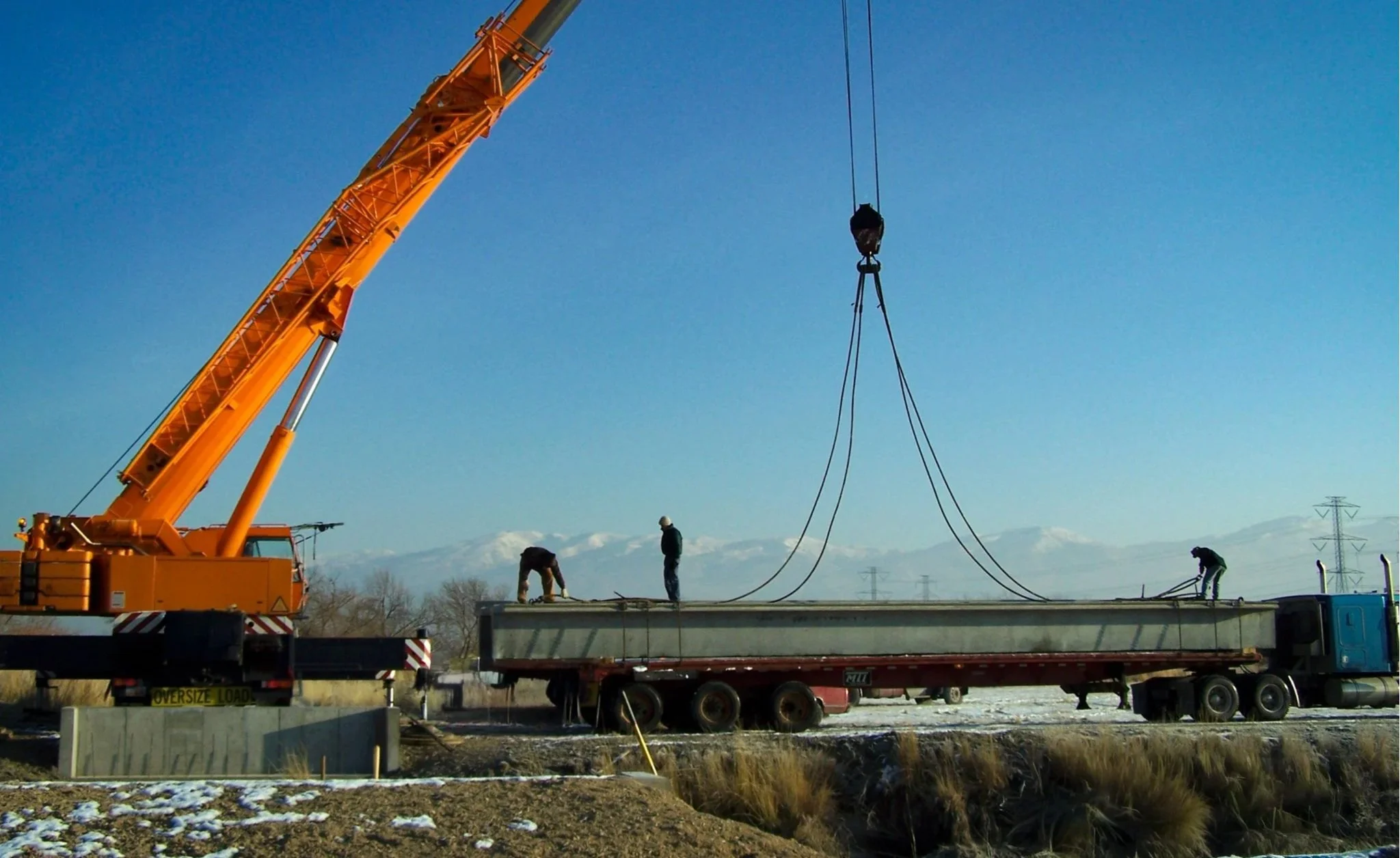 Construction workers on a flatbed truck operate a crane lifting a large concrete beam against a blue sky with distant snow-capped mountains.