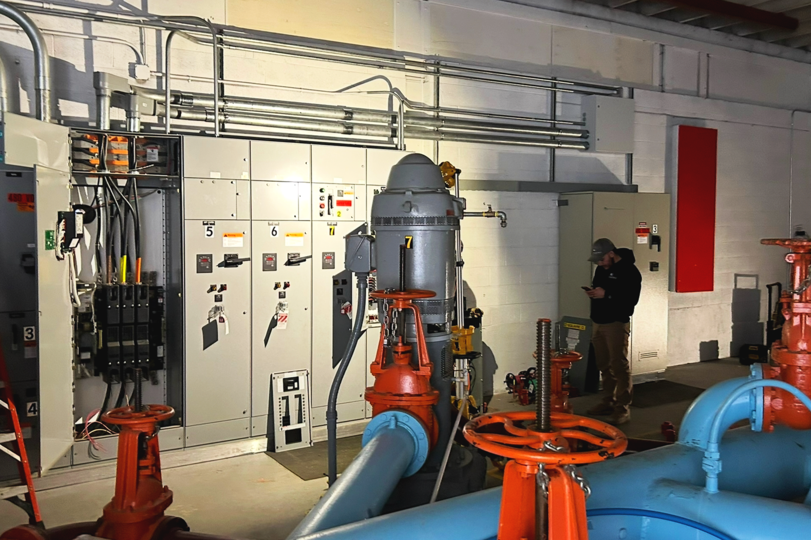 Industrial room with electrical cabinets, pipes, valves, and a person using a mobile phone.