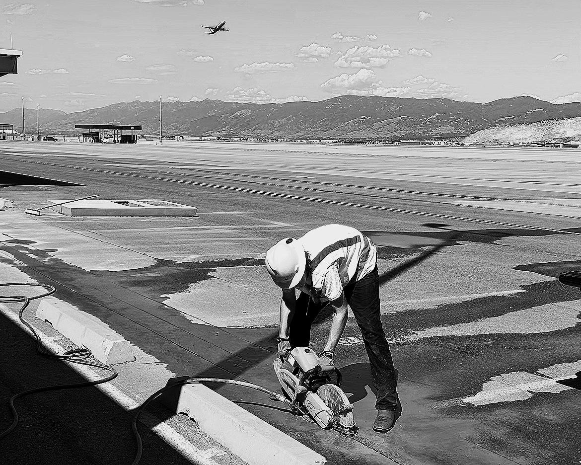 A construction worker wearing a helmet and safety vest using a power saw to cut concrete on a rooftop with an airport runway and mountains in the background, and an airplane flying overhead.
