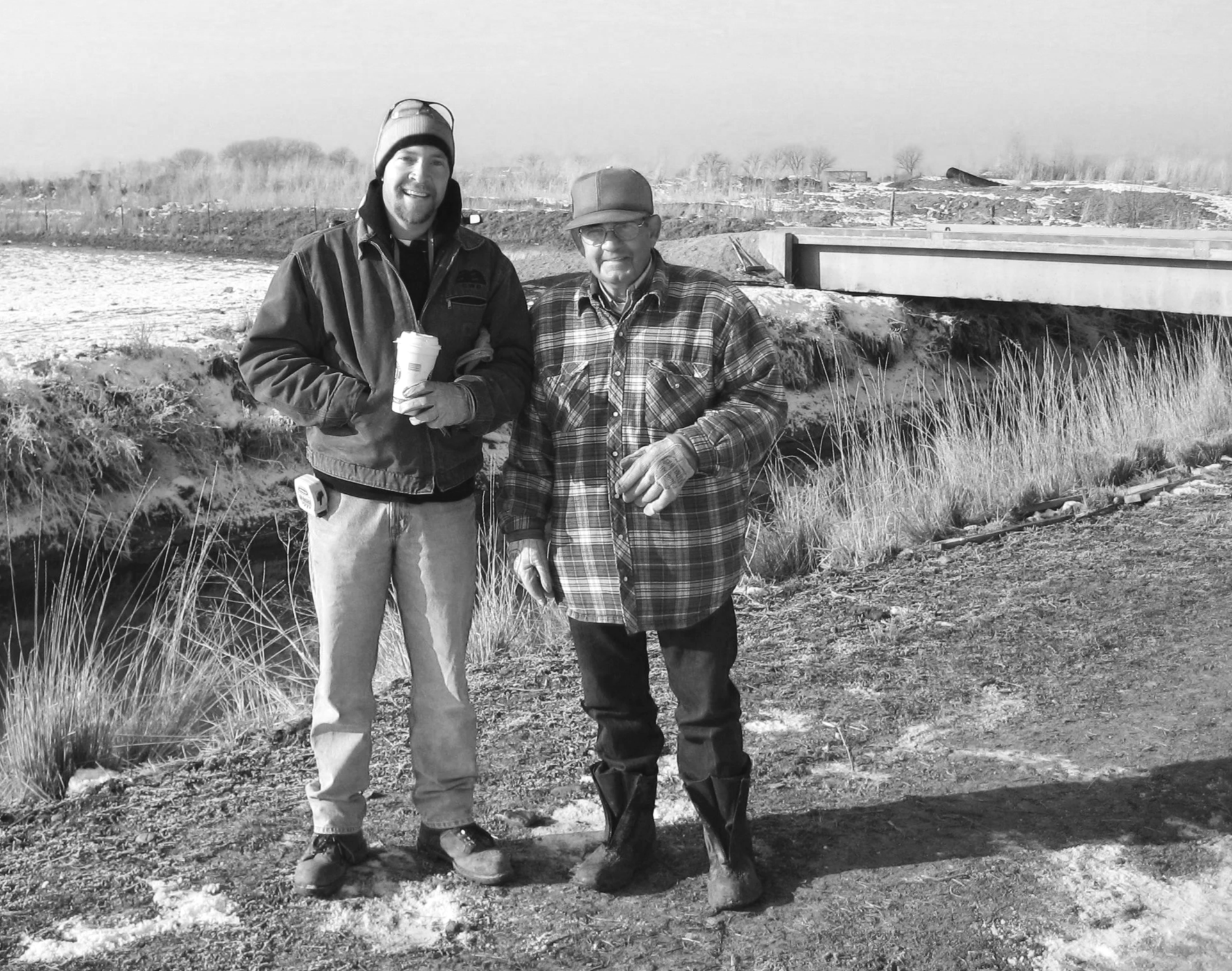 Two men standing outdoors near a small waterway with bare trees and a bridge in the background, in cold weather clothing; one holding a coffee cup.