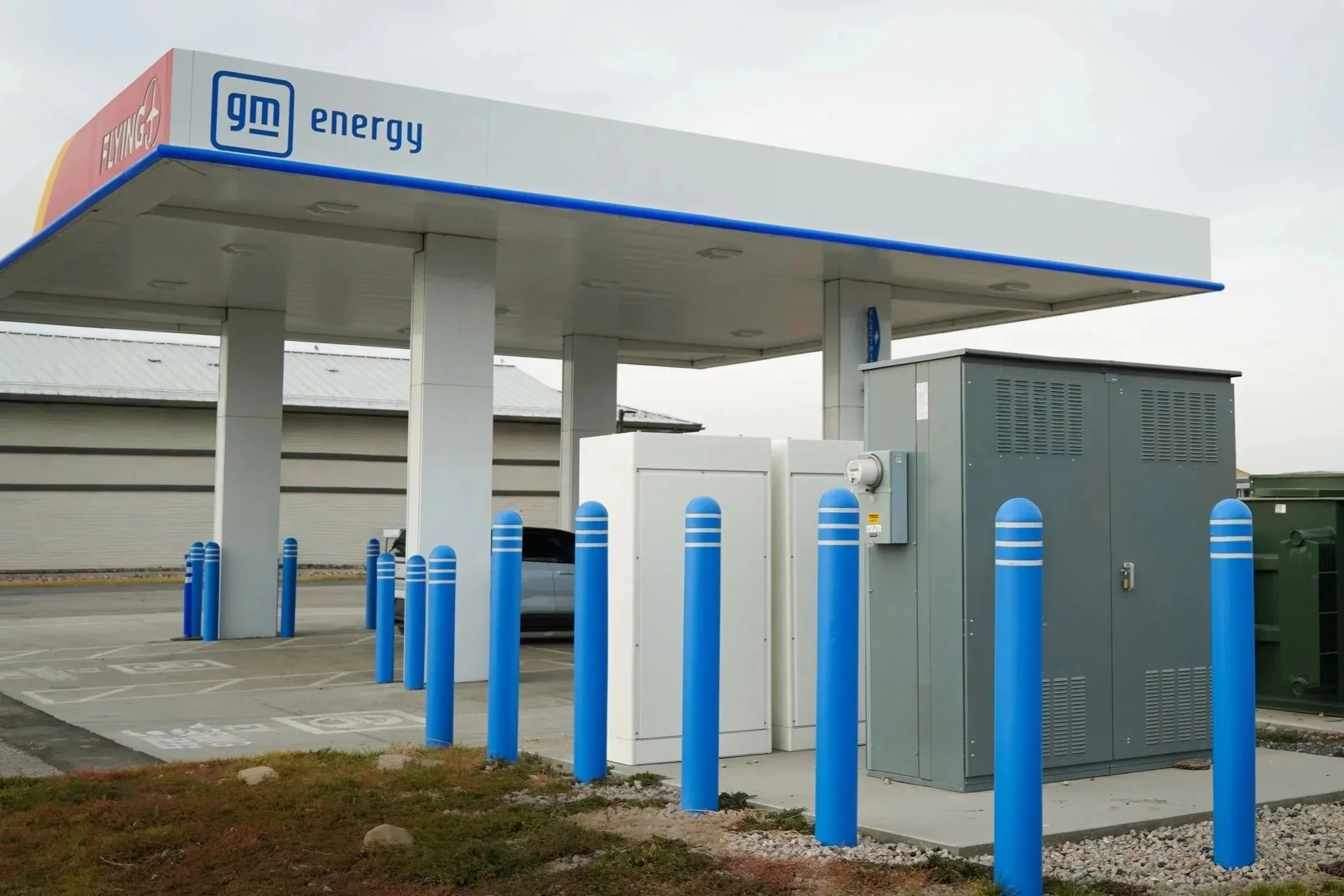 A General Motors energy fueling station with a canopy, blue bollards, electrical boxes, and a parked car.