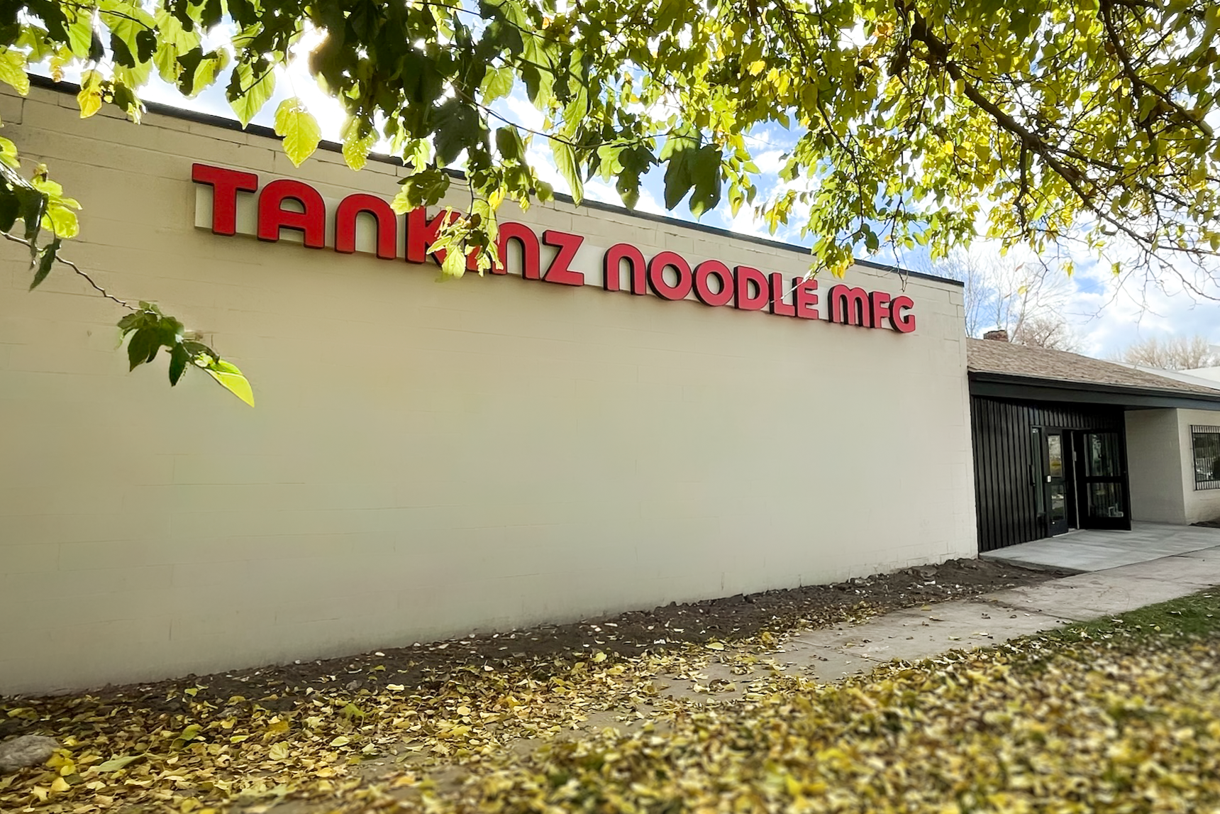 Exterior of a building with a sign that reads 'TANKANZ NOODLE MFG', surrounded by fallen yellow leaves and tree branches.