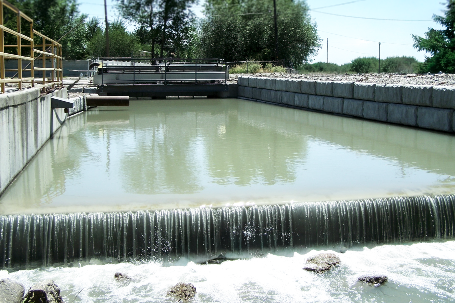 A water treatment plant with a small waterfall, concrete walls on either side, pipes, and a metal gate in the background, surrounded by trees and power lines.