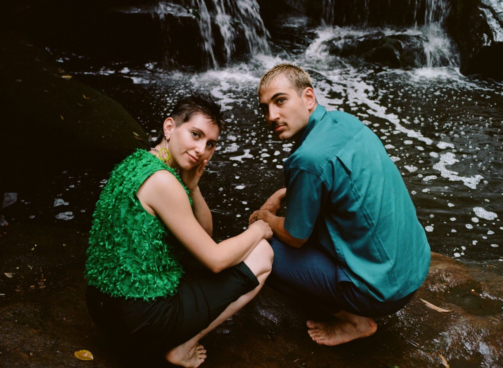 Two people squatting in front of a lake looking back at the camera