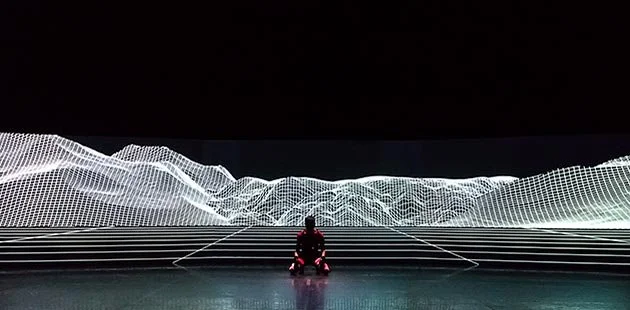 Performer kneeling facing the camera in front of a futuristic background