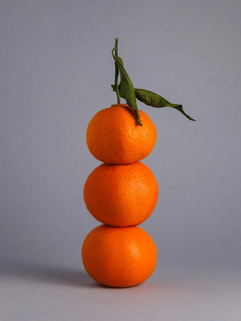 Trois oranges empilées en colonne avec une branche et une feuille au sommet sur un fond gris.