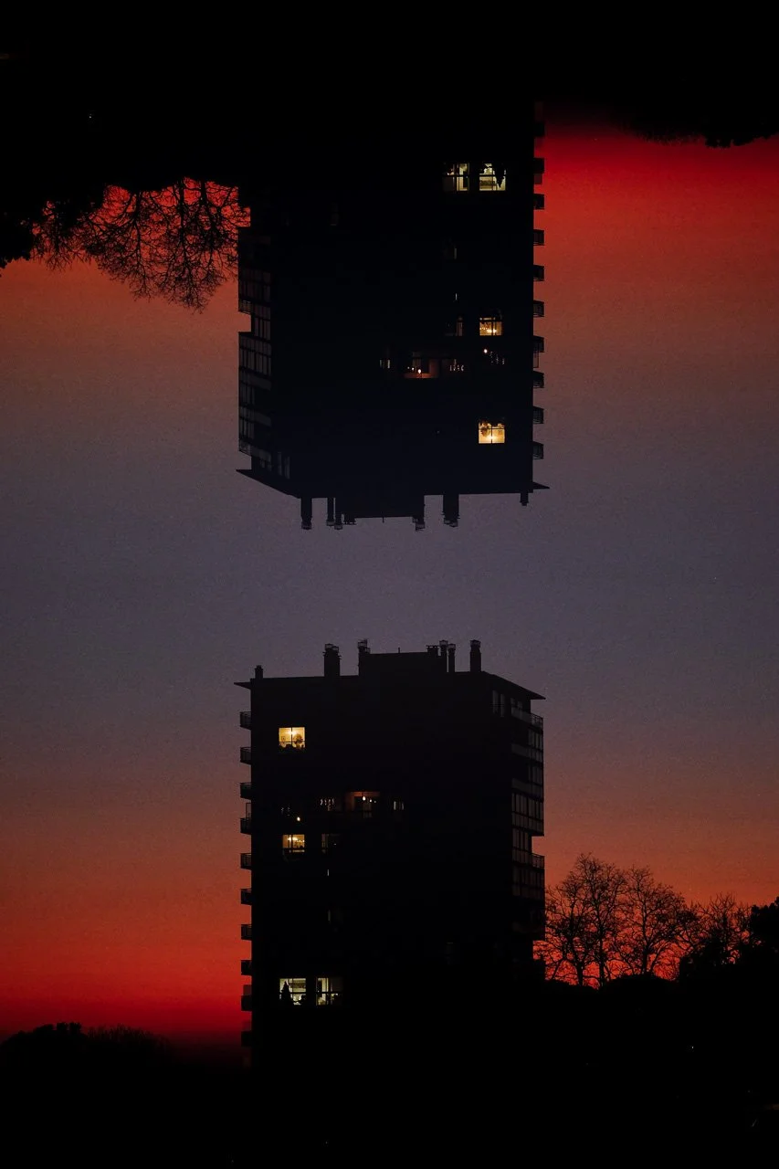 Cliché de deux immeubles résidentiels en contre-jour lors du coucher de soleil, avec des fenêtres éclairées et des arbres silhouettes à l'arrière-plan.