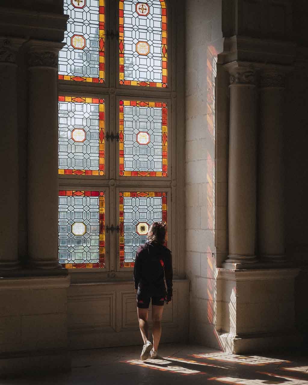 Une personne regarde par une grande fenêtre en vitraux colorés dans une salle avec colonnes du château de Chambord .