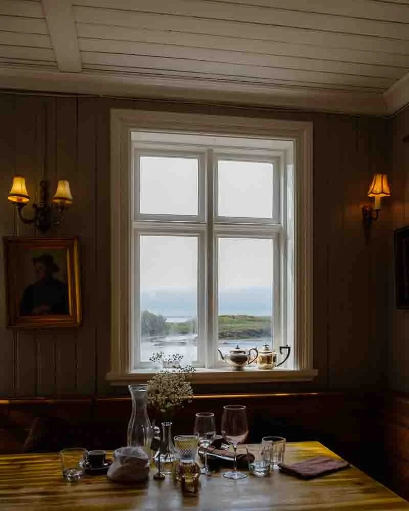 Une table de restaurant en bois avec des verres, une carafe, des assiettes, un bouquet de fleurs, et un napperon. Une fenêtre montre une vue de la campagne et de l'eau en arrière-plan, avec des lampes murales allumées.