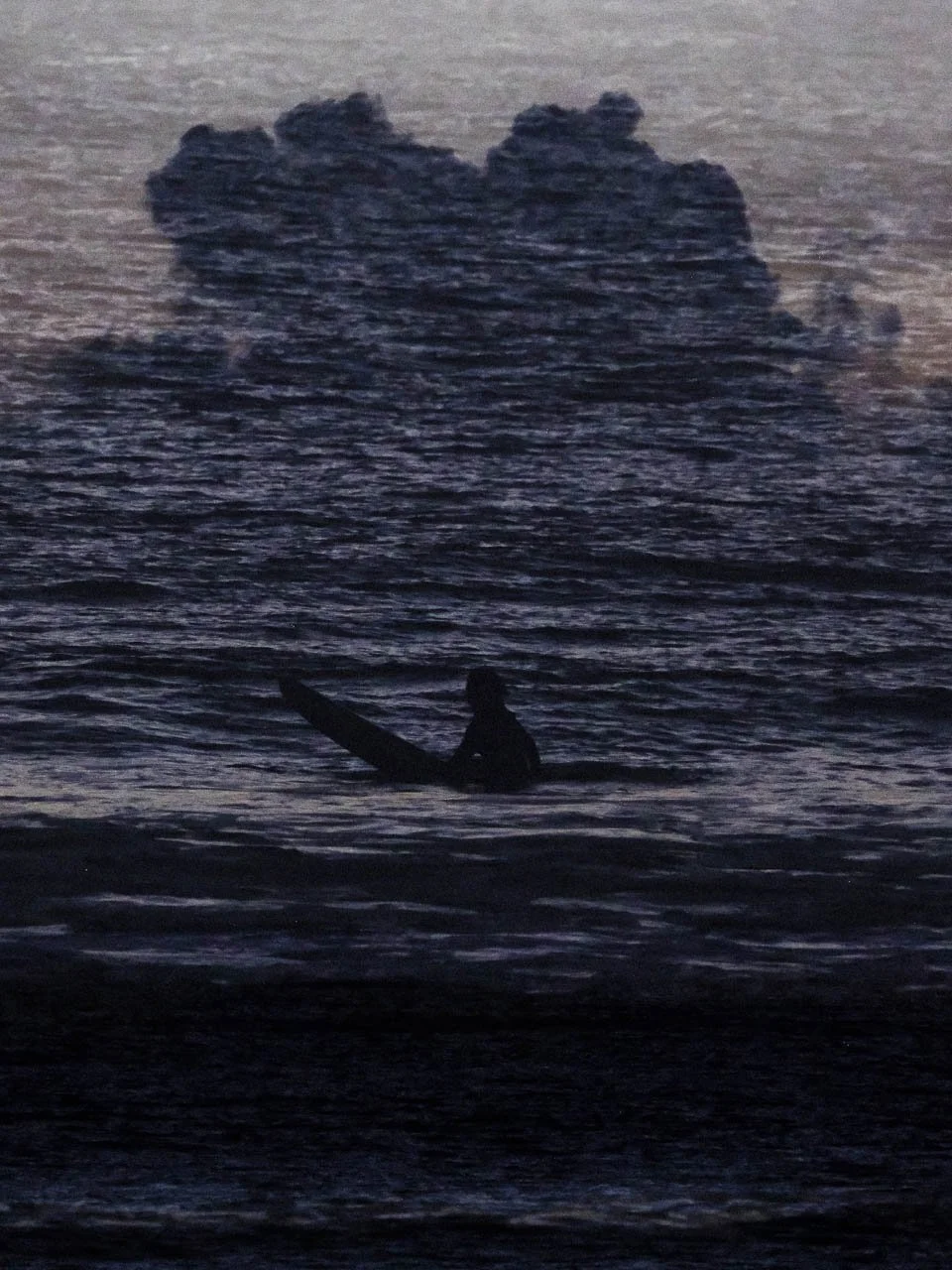 Silhouette d'un surfeur sur une planche dans l'eau, avec un nuage en arrière-plan lors du coucher du soleil.