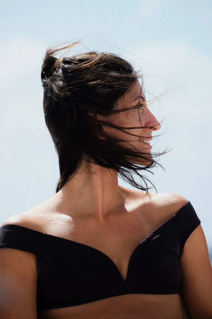Femme souriante avec des cheveux courts et bruns, coiffés avec du vent, portant un haut noir à épaules dénudées, en extérieur par temps ensoleillé.