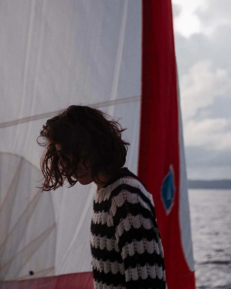 Une femme aux cheveux bouclés et vêtue d'un pull à rayures noirs et blancs, regardant vers le bas, à côté d'une voile de bateau rouge avec un logo bleu, face à la mer sous un ciel nuageux.