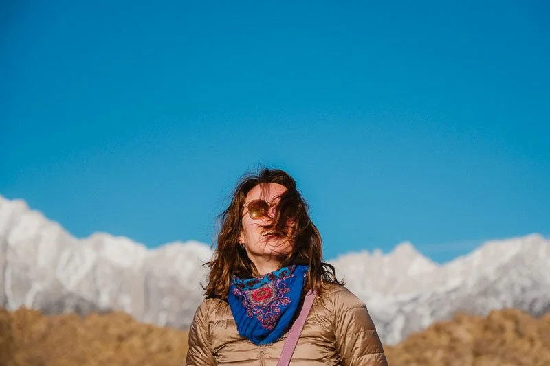 Une femme portant des lunettes de soleil, une écharpe bleue et un manteau beige se tient devant des montagnes enneigées et un ciel bleu clair.