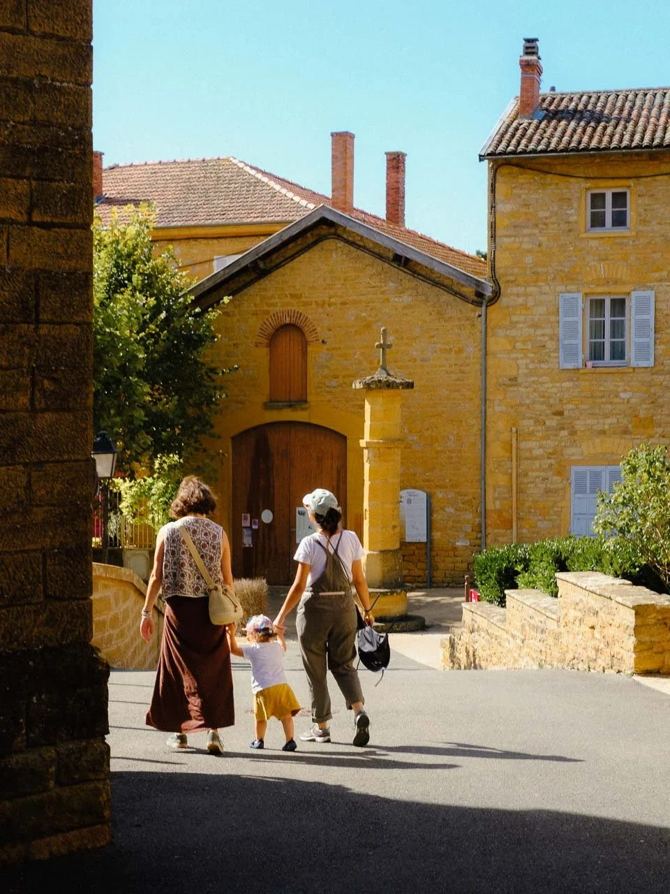 Une famille de trois personnes, deux adultes et un enfant, marchant dans une rue ensoleillée, avec des maisons en pierre de couleur ocre en arrière-plan.