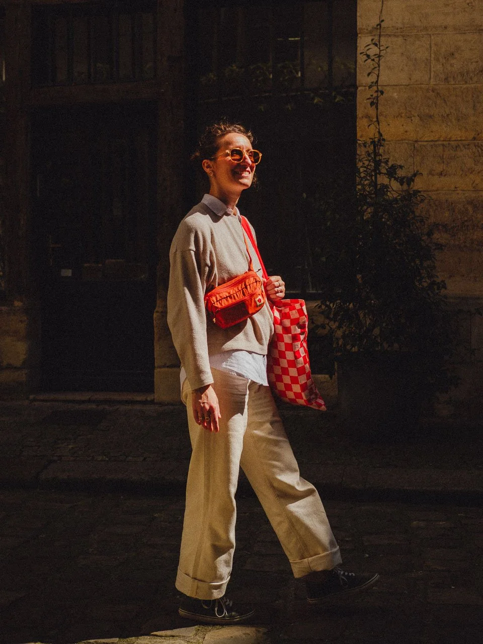 Une femme souriante portant des lunettes orange, une veste beige, un sac rouge, et un sac en filet rouge avec un motif à damier blanc et rouge, marchant dans une rue.