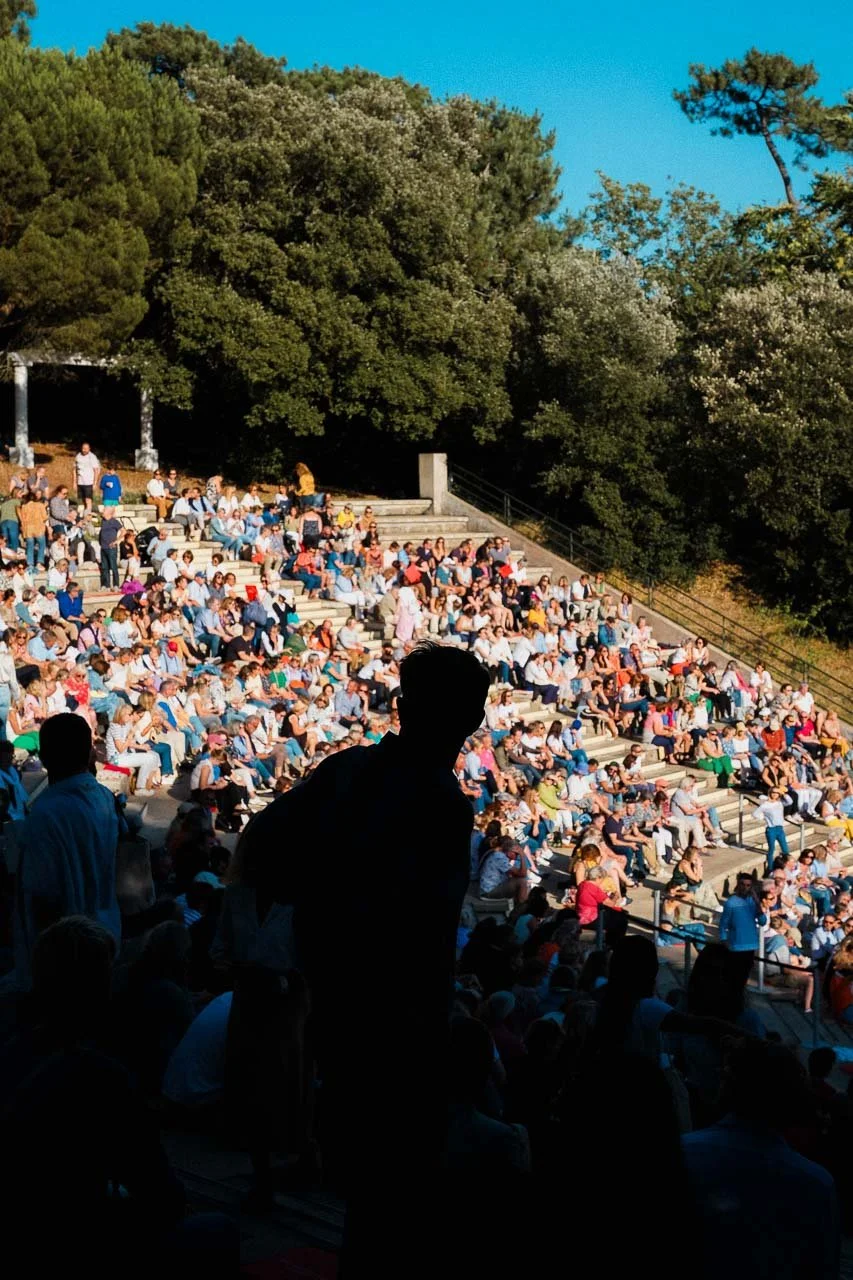 Spectacle en plein air avec une grande foule de spectateurs assis sur des gradins en plein soleil, entourés d'arbres verts et un ciel bleu.