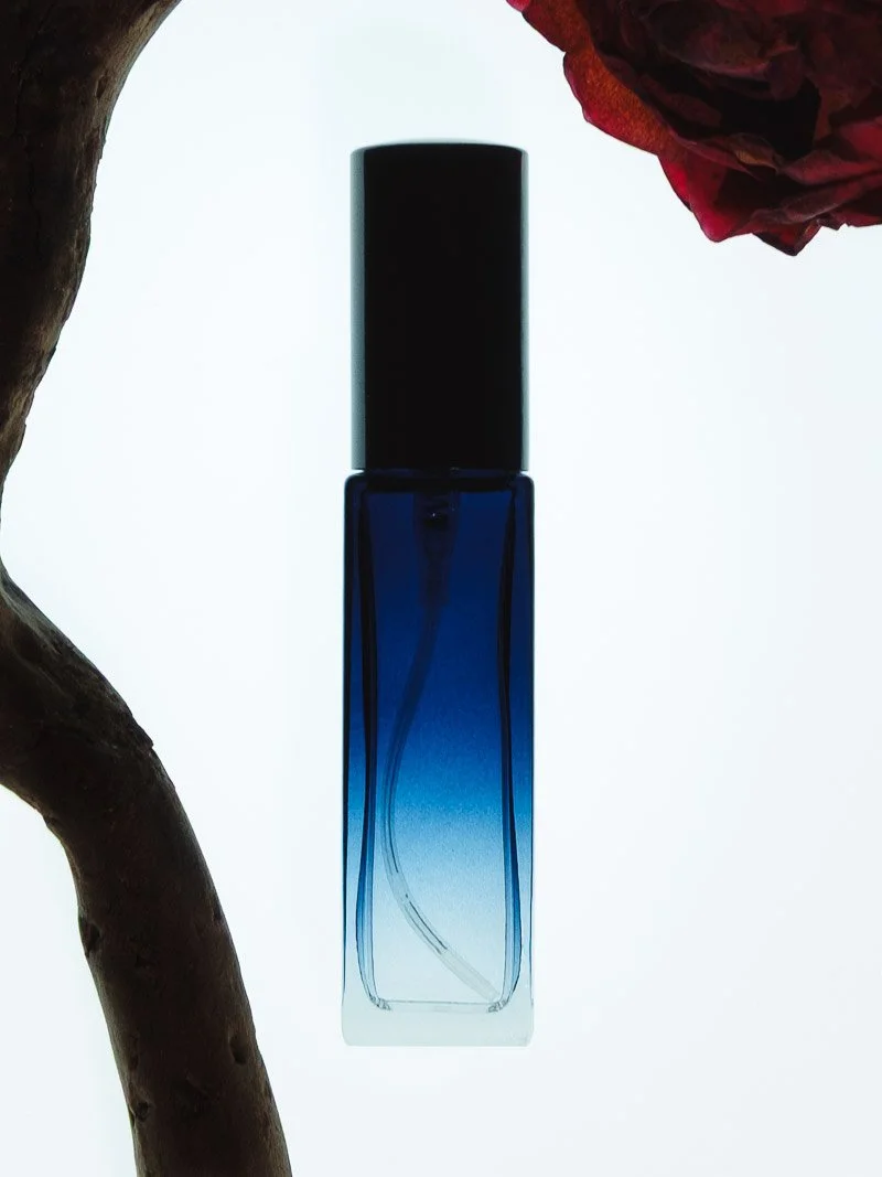 Un flacon de parfum bleu foncé avec un capuchon noir, placé sur un fond lumineux avec des éléments de décoration comme une branche de bois et une fleur rouge.