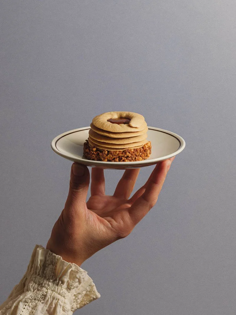 Une main tient une petite assiette avec un biscuit rond, plusieurs cookies ou biscuits ronds empilés dessus, posés sur un biscuit croquant ou granola.