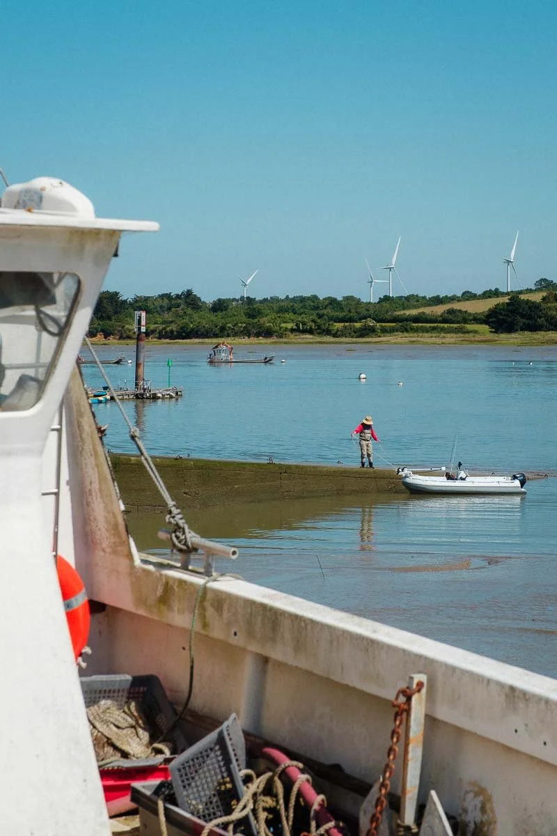 Vue du littoral avec un bateau au premier plan, une personne en train de pêcher, et des éoliennes au loin sous un ciel dégagé.