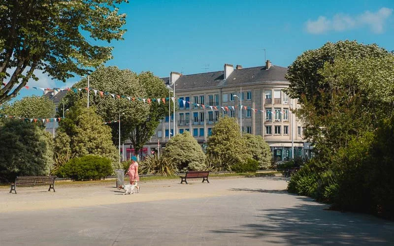 Place de la mairie de Saint-Nazaiez avec des bancs, des arbres et des décorations de drapeaux colorés, avec un bâtiment résidentiel en arrière-plan et un ciel bleu clair.