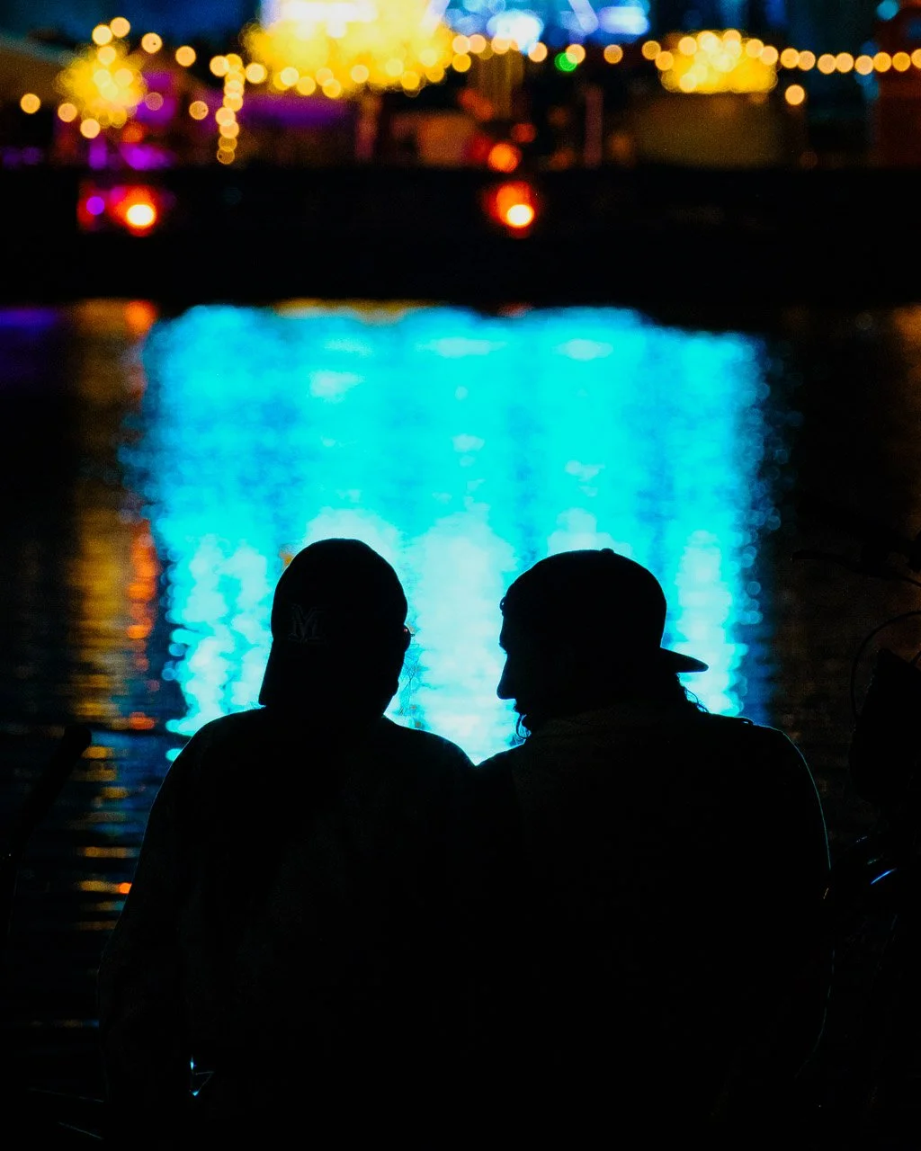 Deux personnes en silhouette assises près de l'eau, avec des lumières colorées floues en arrière-plan, lors d'un festival.
