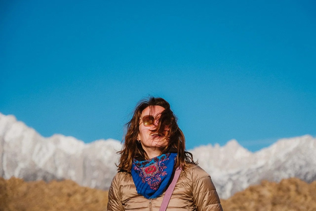Femme portant des lunettes de soleil, un foulard bleu et une veste beige, debout devant une montagne enneigée et un ciel bleu clair aux USA
.