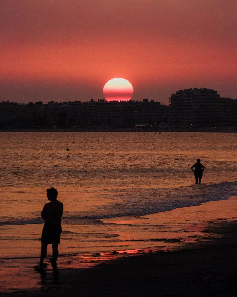 Coucher de soleil sur la mer avec deux enfants en silhouette sur la plage et un horizon urbain en arrière-plan.