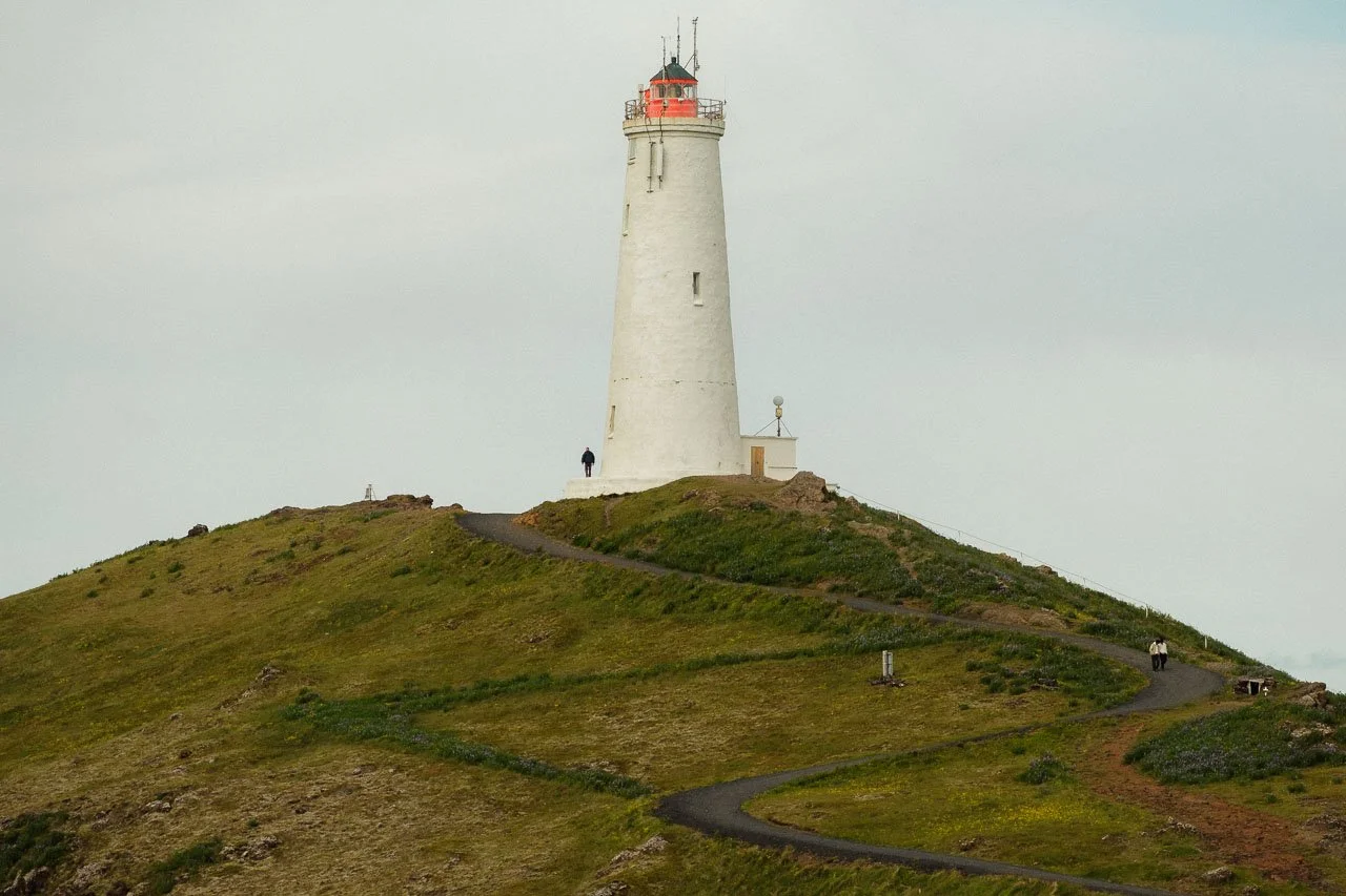 Phare blanc sur une colline verte avec un chemin sinueux, avec quelques personnes autour.