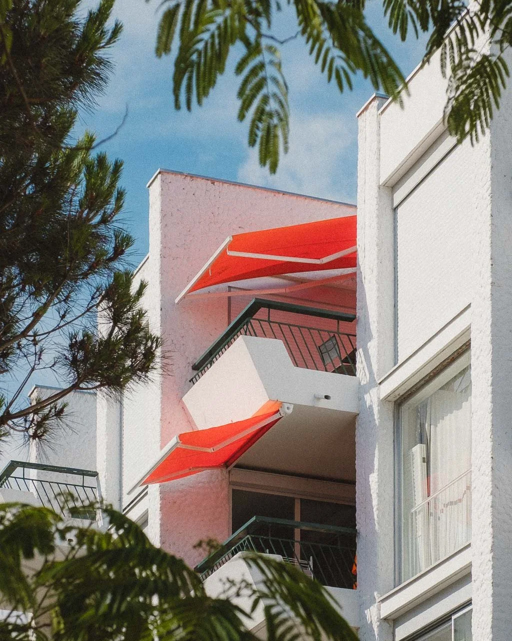 Balcons avec stores orange sur un immeuble blanc, entouré de feuilles d'arbres, sous un ciel bleu à la baule.