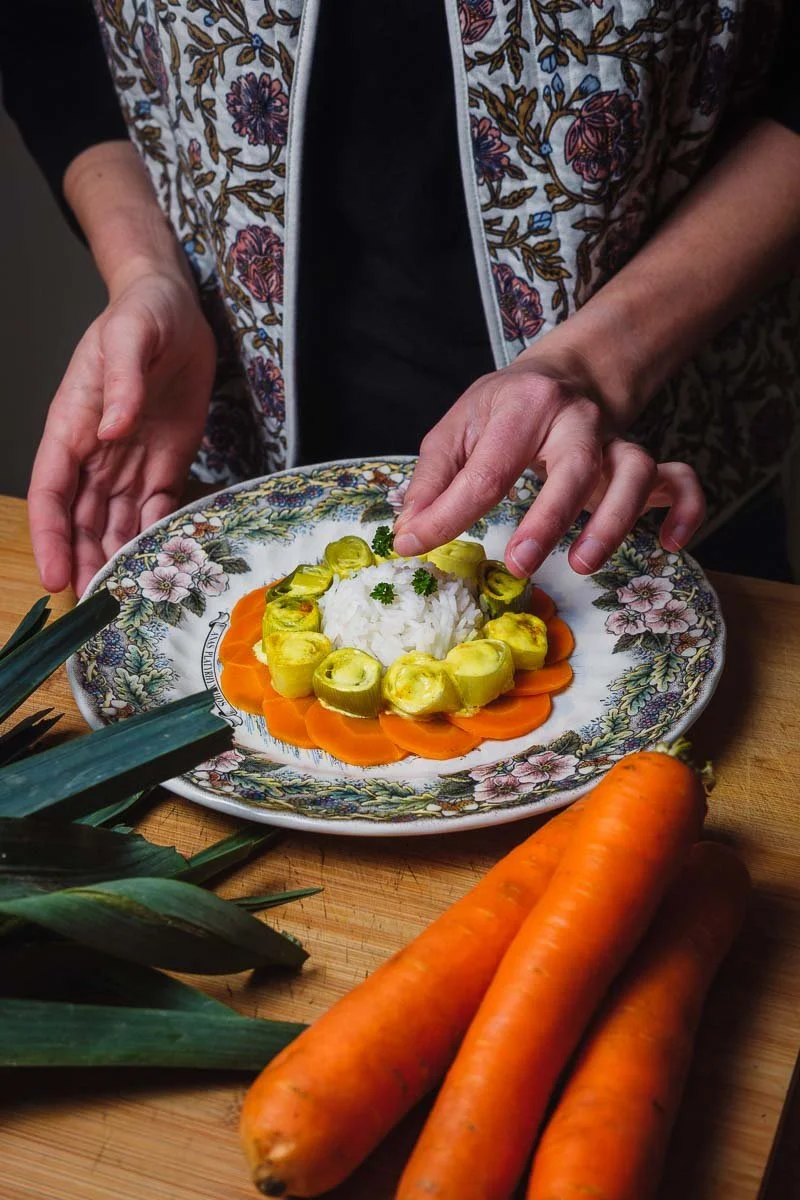 Une personne prépare un plat avec du riz blanc au centre, entouré de tranches de carotte et d'ensembles de petits légumes verts sur une assiette décorative. Il y a également des carottes entières et des feuilles d'ananas sur la table.