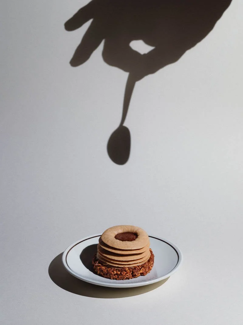 Une assiette avec plusieurs biscuits ronds, empilés et saupoudrés de cacao, avec une base croquante. Une ombre de main portant une cuillère tombe au-dessus de l'assiette, créant un jeu d'ombre et de lumière.