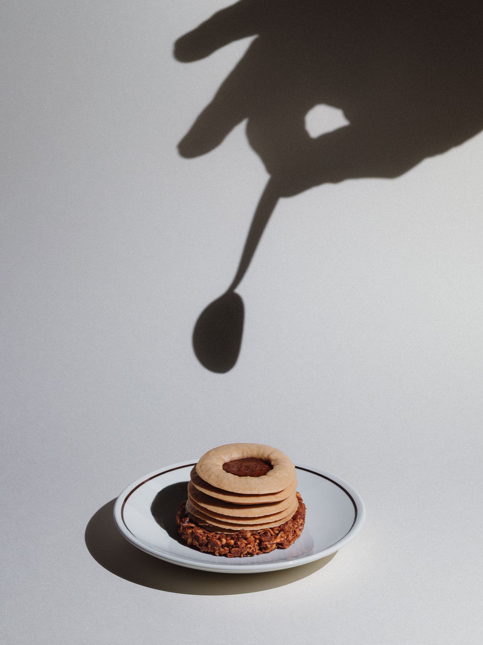 Un gâteau en forme de tour avec plusieurs couches de biscuits et une base en crumble, placé sur une assiette blanche. La scène est éclairée pour créer une ombre d'une main tenant une cuillère qui descend vers le gâteau.
