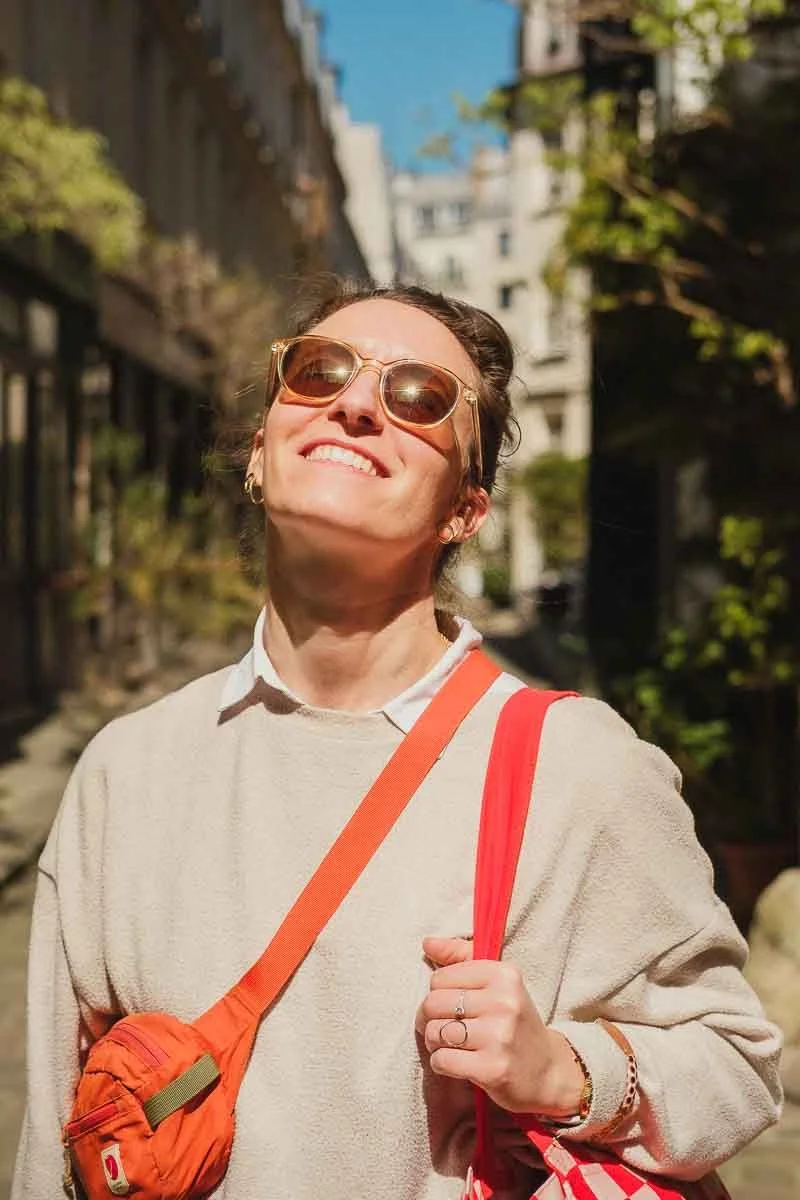 Une femme souriante portant des lunettes de soleil, un sweat beige, un sac orange et une bandoulière rouge, se tenant à l'extérieur en pleine journée ensoleillée, avec des bâtiments en arrière-plan