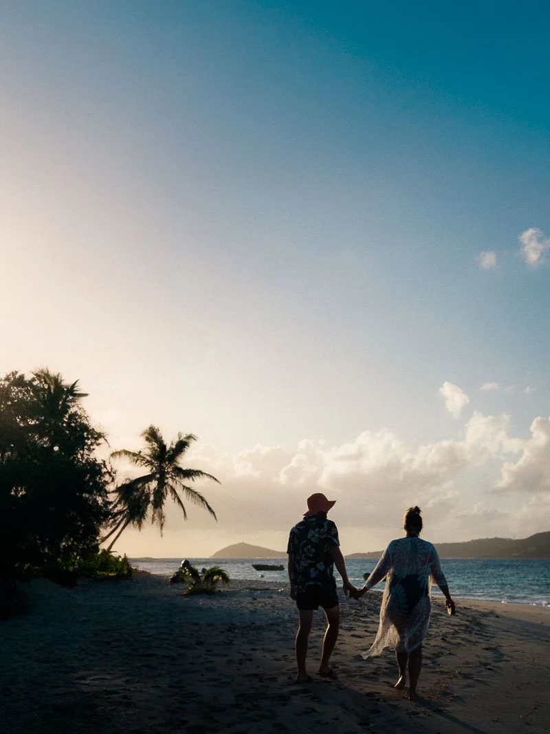 Un couple marche main dans la main sur une plage de sable, avec des palmiers et la mer à l'arrière-plan, au coucher du soleil.