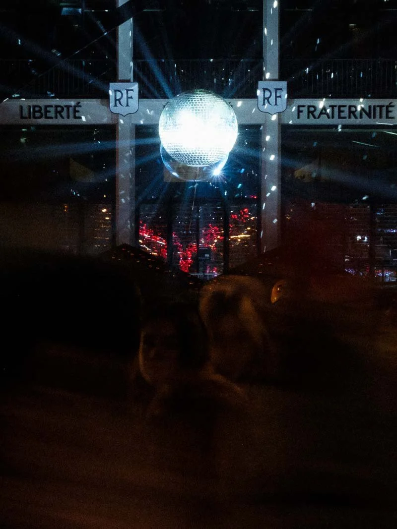 Fête de noël sur la place de la mairie de Saint-Nazaire, drapeaux français avec les mots "Liberté" et "Fraternité" et les lettres "RF". Éclairage coloré et ambiance sombre.