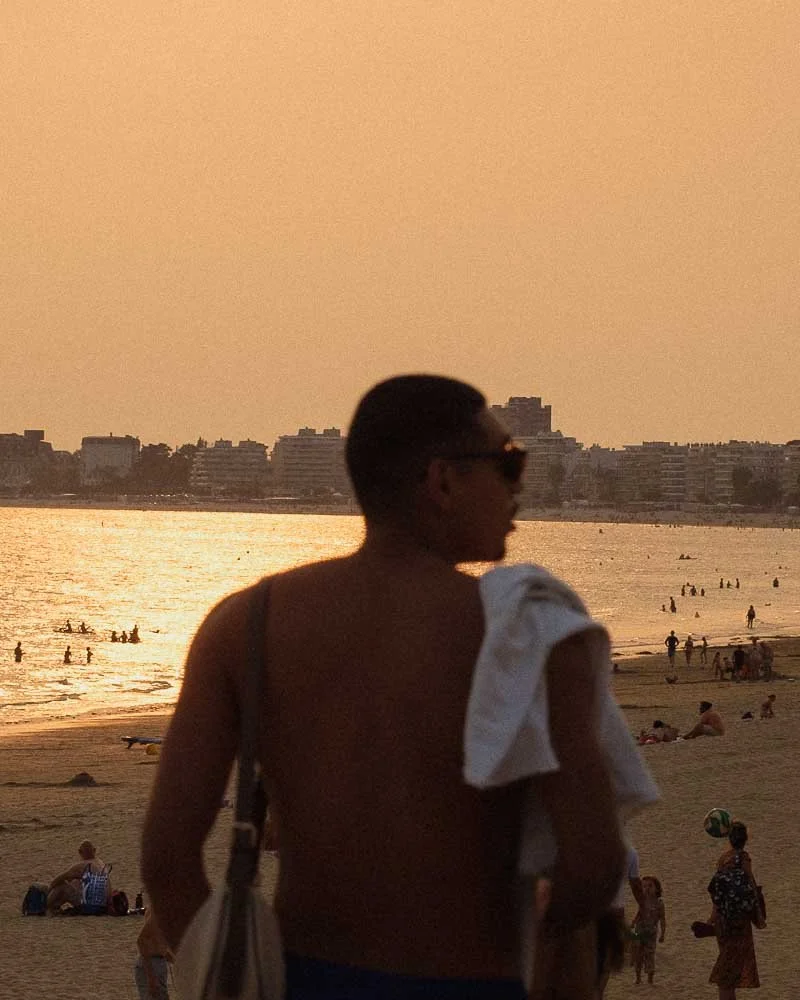Un homme debout sur la plage de pornchet au coucher du soleil, portant des lunettes de soleil et un vêtement blanc sur l'épaule, avec une ville en arrière-plan.