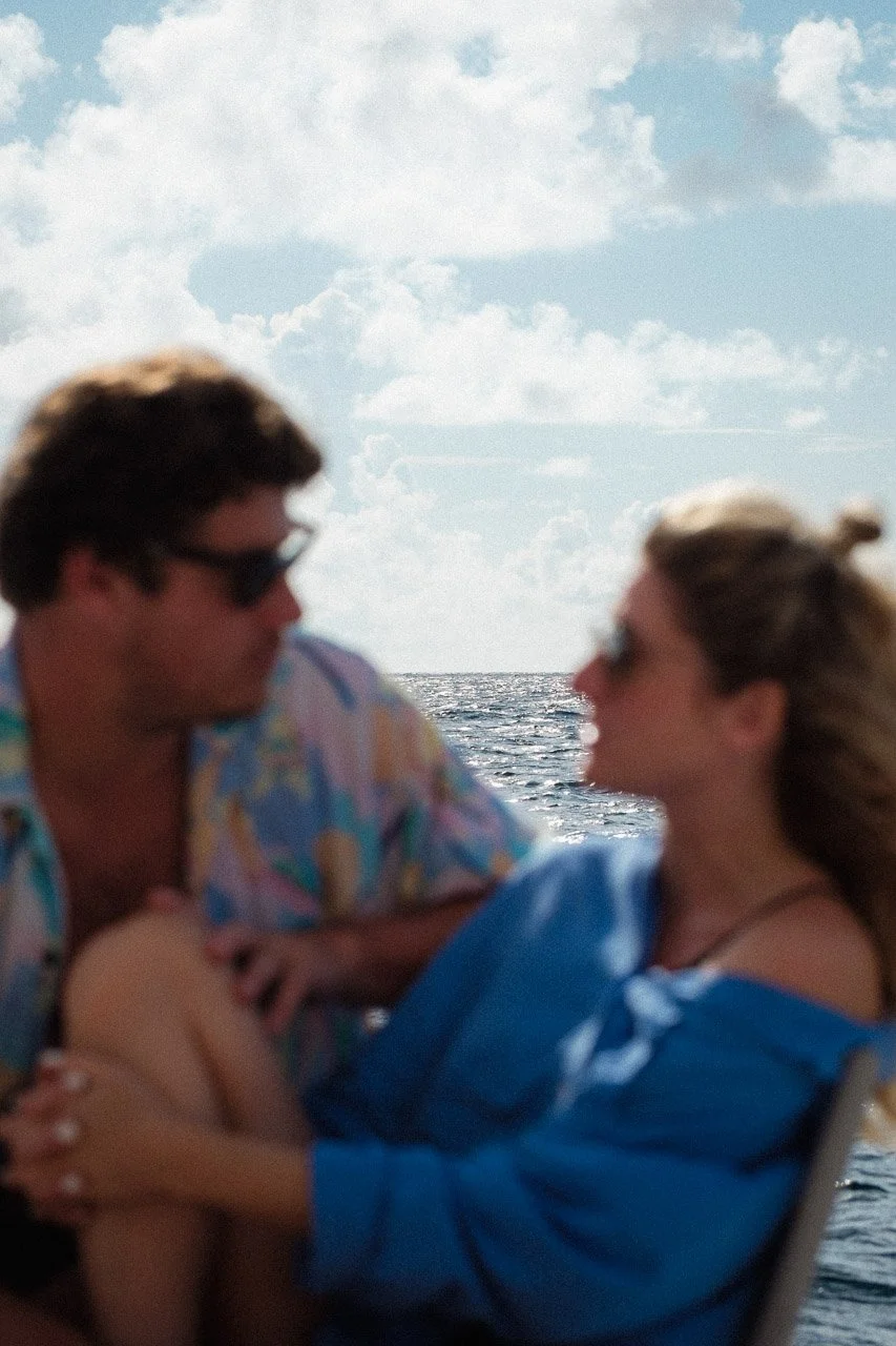 Deux personnes assises près de la mer, échangeant un regard, avec un ciel bleu et des nuages en toile de fond.