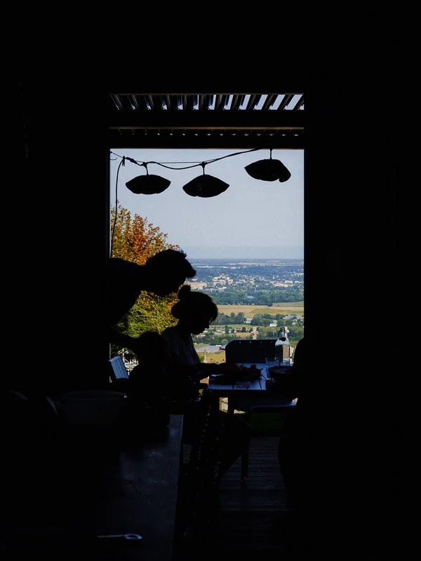 Silhouettes d'une famille à une table à l'intérieur d'une cabane en bois, regardant un paysage avec des champs et un ciel clair, avec des lampes suspendues au plafond.