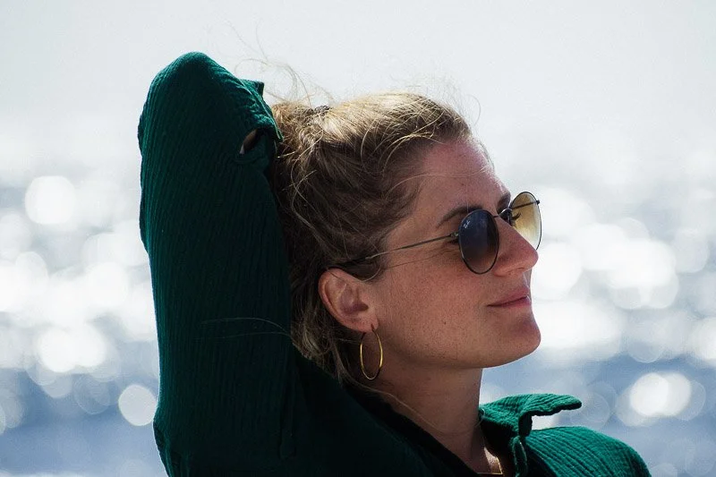 Une femme portant des lunettes de soleil et un earring en or, détendue, avec le soleil et la mer en arrière-plan.