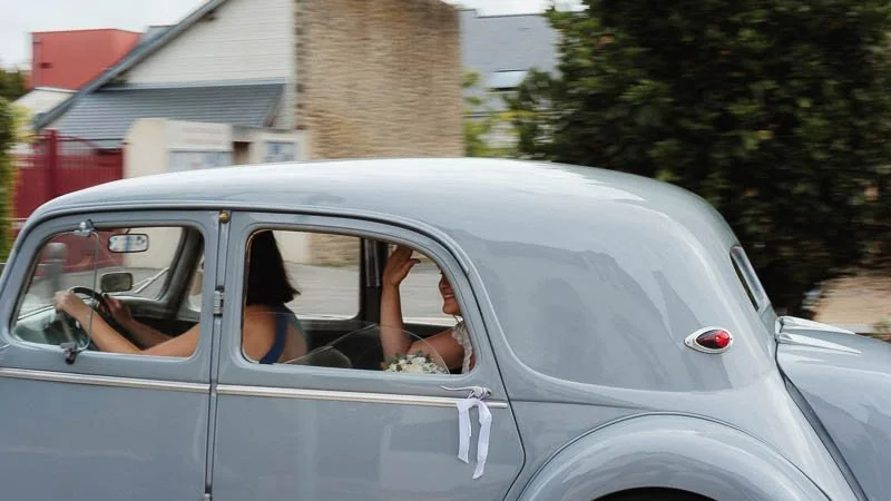 Voiture ancienne gris clair avec les mariés à l'intérieur, conduisant dans une rue résidentielle.