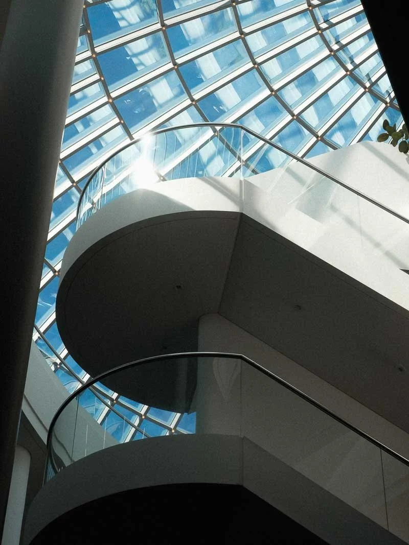 L'intérieur d'un bâtiment moderne avec un plafond en verre en forme de dôme, montrant deux niveaux avec des balustrades en verre et un escalier tournant.