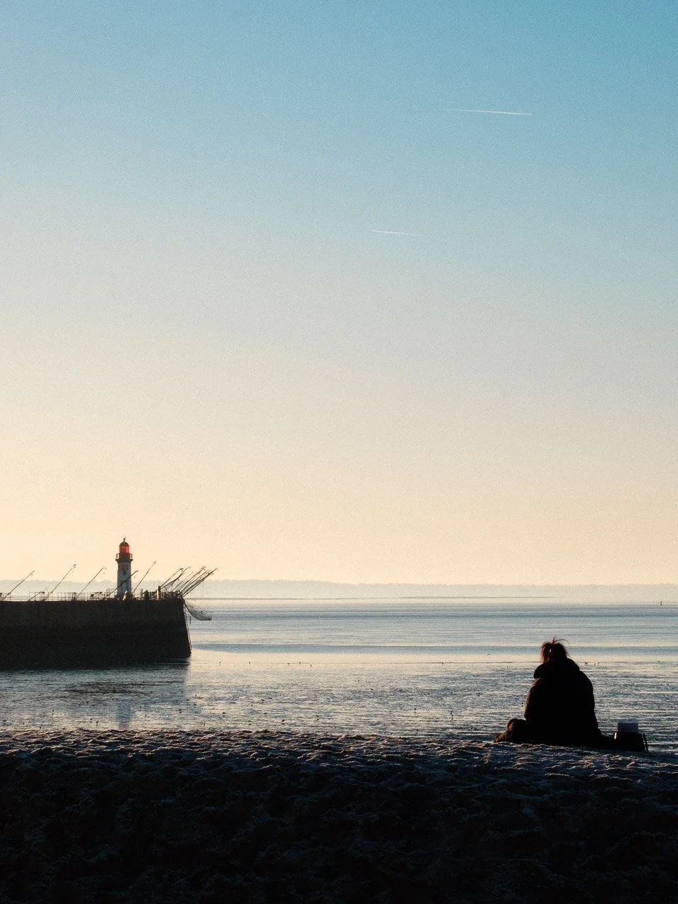Personne assise sur la plage regardant la mer au lever du soleil, avec un phare et un quai à gauche.