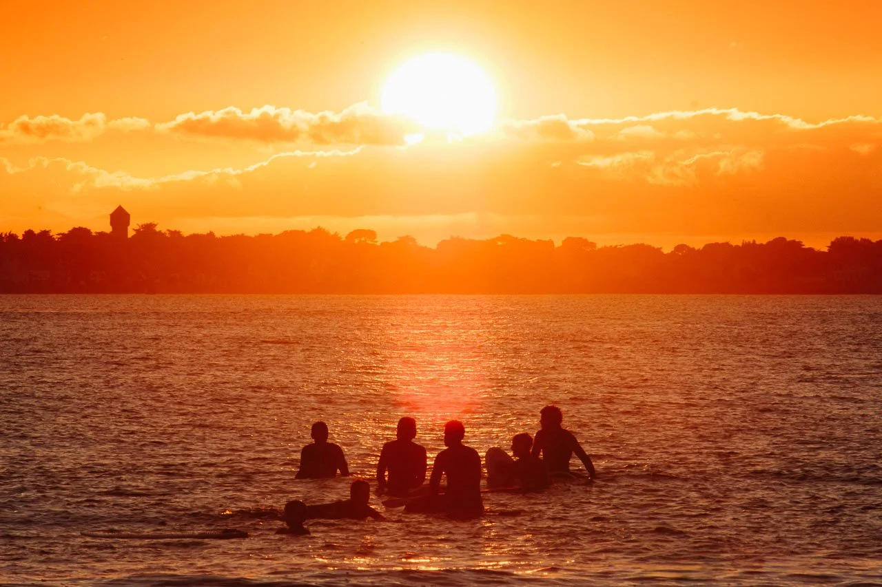 Groupe de surfeur dans une mer au coucher du soleil avec un ciel orange et un paysage en arrière-plan.