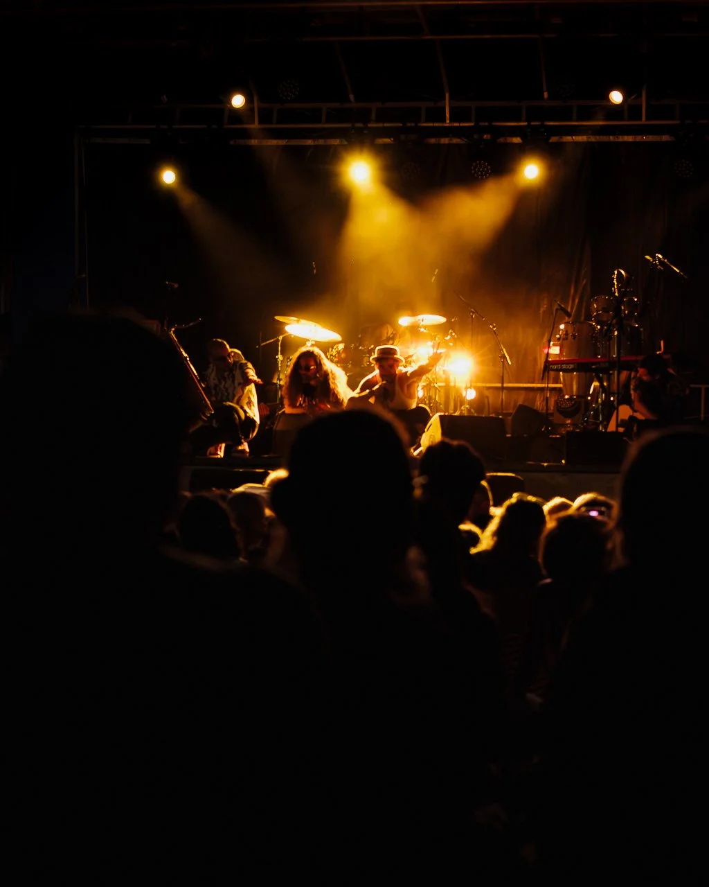 Une scène de concert avec un groupe sur scène sous un éclairage chaud, des instruments de musique et un public regardant le spectacle dans une ambiance sombre.