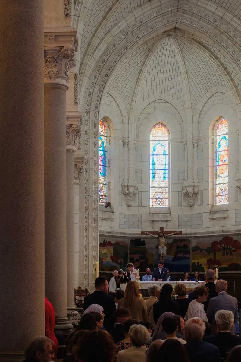 Une cérémonie de mariage religieuse dans une église avec des personnes assises et debout, un autel avec une croix et un crucifix, des vitraux colorés et une architecture en pierre blanche avec des arches.