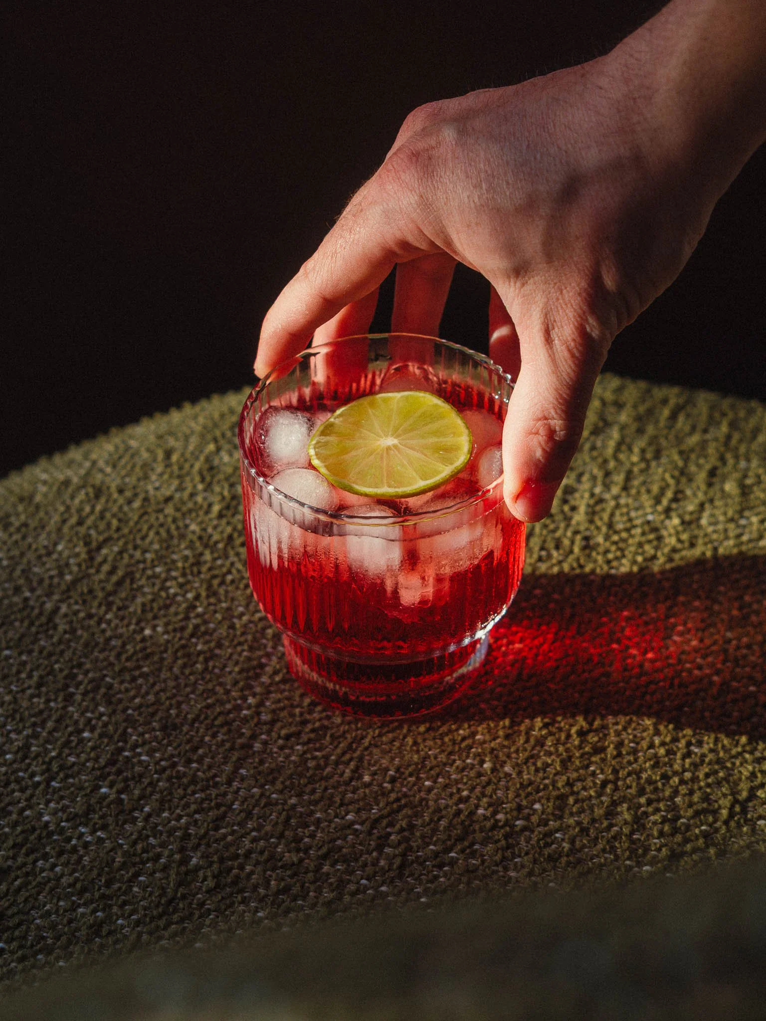 Main tenant un verre à moitié rempli d'une boisson rouge avec des glaçons et une tranche de citron vert, posé sur une surface texturée, fond sombre.