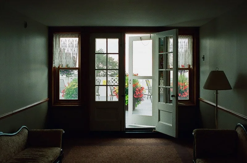 Intérieur d'une pièce avec fenêtre et porte ouverte menant à un balcon avec fleurs et mobilier.
